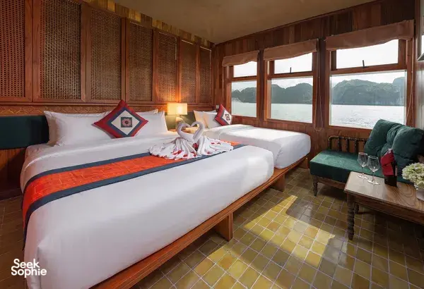 Wooden luxury cabin with a made-up double bed, pillows and side sofa, overlooking calm waters from Cat Ba Ocean Cruises