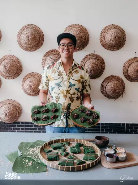 Benny from TempeMan smiling in a Bali kitchen, holding trays of banana-leaf tempe cookies ready to serve