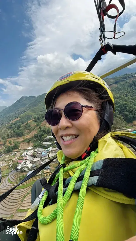 Smiling paragliding passenger in a yellow helmet and sunglasses looks out over green terraces and a river valley under blue sky in Sapa