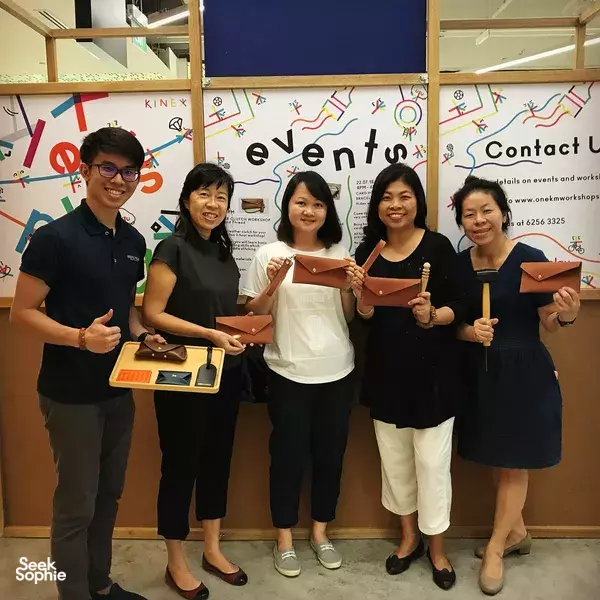 Smiling group holding brown leather clutches at Hides and Thread in Singapore in front of an events wall for workshops in Singapore