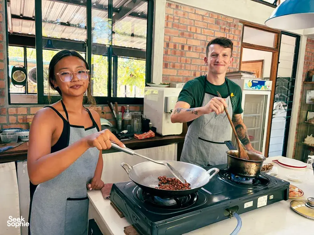 Cooking Malaysian comfort food with two people stirring a wok on a stovetop inside Kebun Lidstari’s farmhouse kitchen, Langkawi