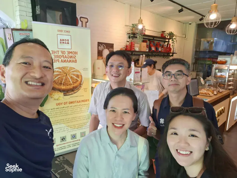 Smiling group indoors by a bakery counter in Katong-Joo Chiat during a real-life Monopoly game in Singapore, team building Singapore