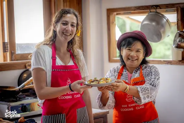 Two women in red aprons smiling while holding plated dishes at the end of the Mae Kampong forage-to-table cooking class in Chiang Mai