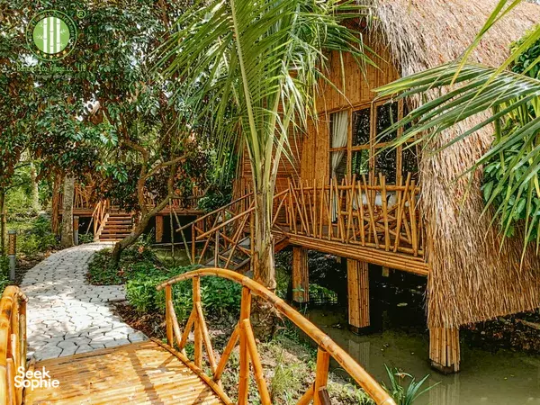 Stilted bamboo deck beside a calm canal at Bamboo Eco Village, with a thatched cabin and tropical garden around the Mekong Delta