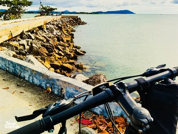 Bike handlebars on a Langkawi coastal route beside a rocky breakwater, with calm sea stretching to distant islands