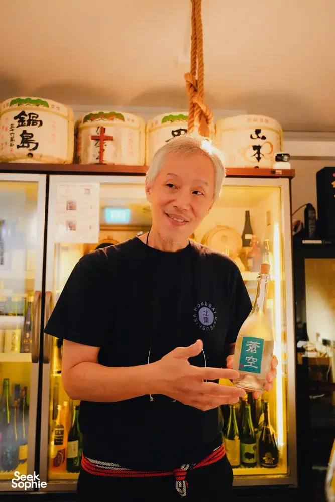Host SakeMama welcoming guests at a rare sake tasting in Singapore, standing by a cabinet of bottles and labels