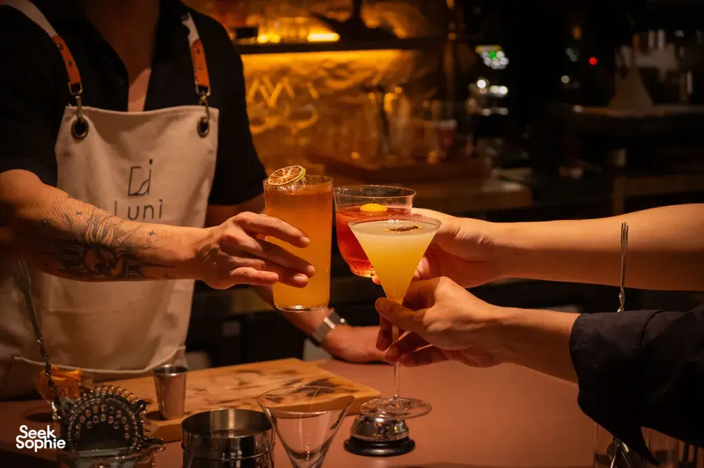 Hands raise two by-the-glass cocktails with citrus garnishes for a toast at an intimate Singapore cocktail bar.