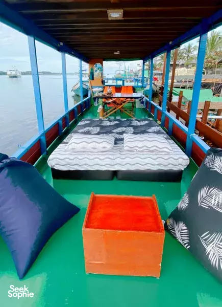 Pillows and a wooden storage box on a small boat deck-bed, with calm river water and other boats visible ahead in Tanjung Puting