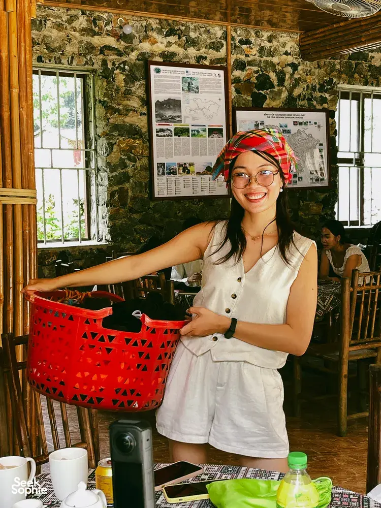 Smiling Ha Giang Expeditions host in a rustic stone room, standing by a table with travel materials and a red basket