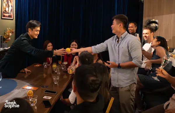 Audience gathered around a magician performing a tabletop trick at The Magic Bar in Singapore, with cocktails and cards on tables