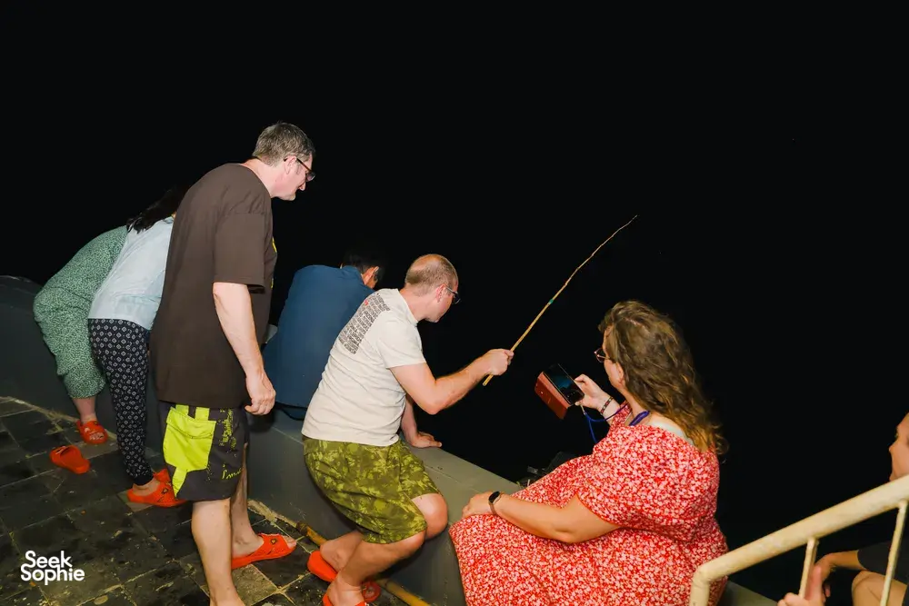 Guests on a night-time cruise deck pulling up a netted catch from the water with a long-handled tool