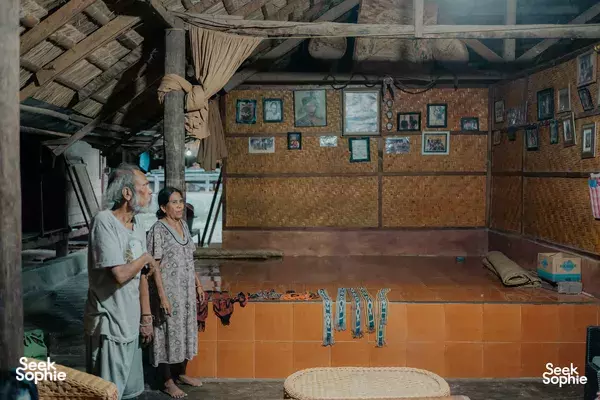 Two travellers standing in a rustic indoor room on Sumba, with tiled flooring, framed pictures along the walls, and a low bed