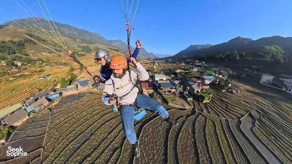 Helmeted paraglider flying above patchwork terraced fields and rooftops, legs lifted, with Sapa Paragliding over Sapa