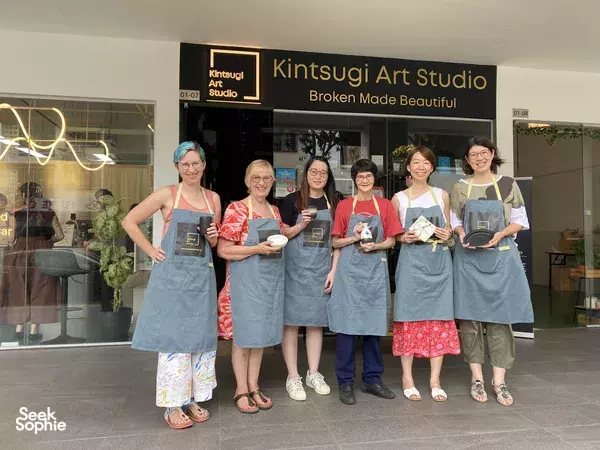 Group of participants in aprons posing outside Kintsugi Art Studio with their mended art pieces, during workshops in Singapore