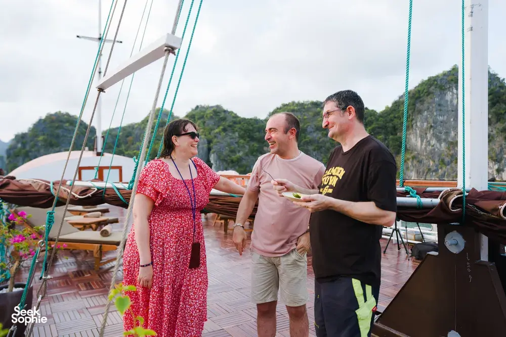 Travellers chatting on the deck of a boutique wooden cruise in Cat Ba National Park, with limestone cliffs across the bay