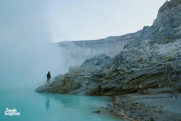 Hiker standing on a foggy ledge beside a turquoise lagoon near jagged cliffs, with distant clouds in the valley