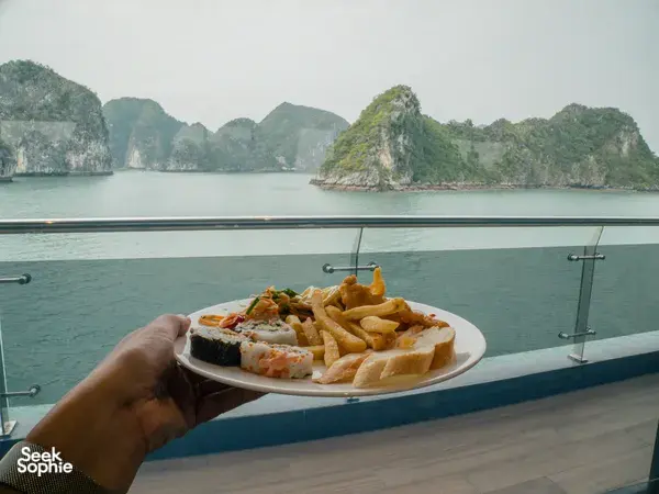 Hand holding a plate of appetisers by the deck rail, with misty karst islands across Lan Ha Bay during a Tulip Cruise sailing