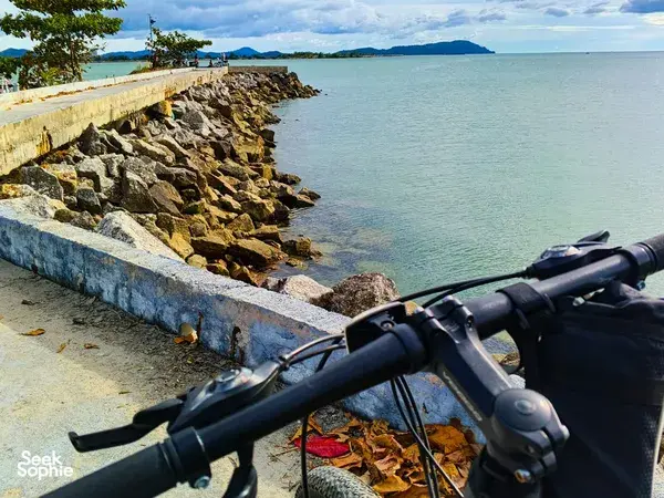 Bike handlebars rest on a rocky seawall while calm water and islands stretch along the coast of Langkawi