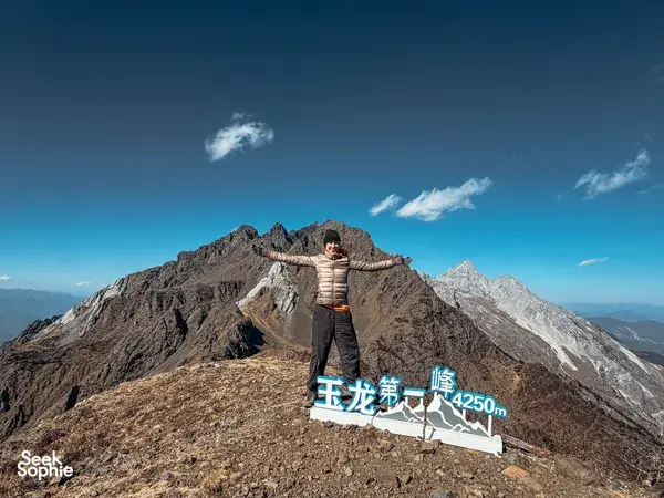 Jade Dragon Snow Mountain hike from Lijiang: hiker posing on a rocky summit ridge beside a 4,250m marker, host-led by The Bivou Lijiang