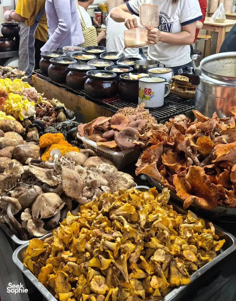 Trays of cooked meats and mushrooms at a Lijiang market stall, with sizzling pots behind and diners with cups