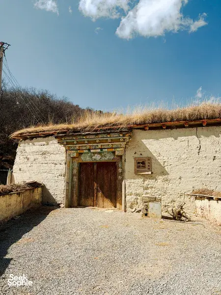 Whitewashed Tibetan homestead doorway at Ferme Liotard in Shangri-La, with carved lintel and tall grass under blue sky