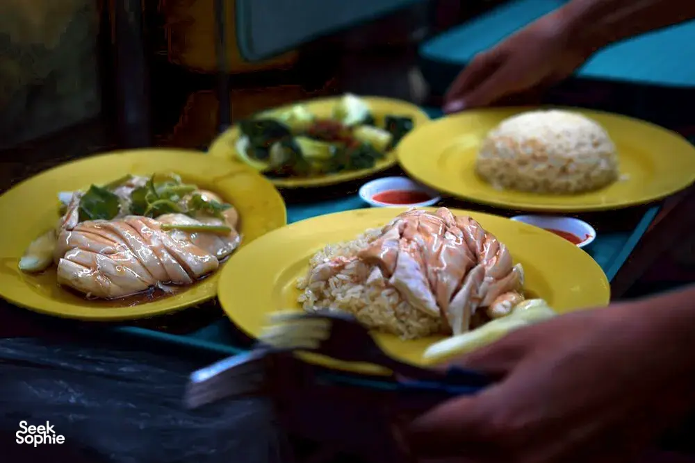 Hands carrying yellow plates of hawker food at a Singapore stall, showcasing Singapore hidden gems from the city’s hawker culture