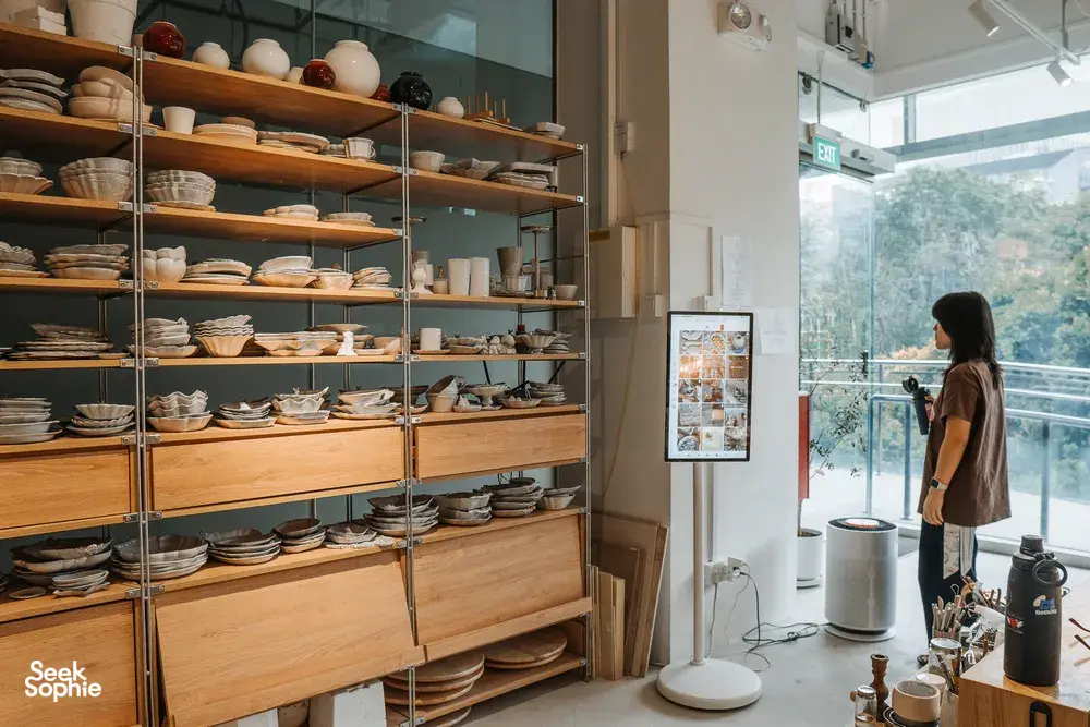 Rows of handmade ceramic plates on tall shelving in Ina Atelier’s Singapore studio, with a person standing by the window