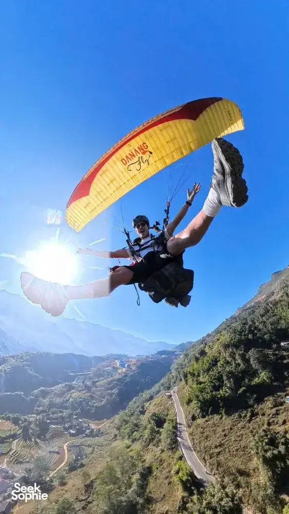 Paraglider rider in mid-air with a yellow canopy framed against bright blue sky and a winding mountain valley road, Sapa