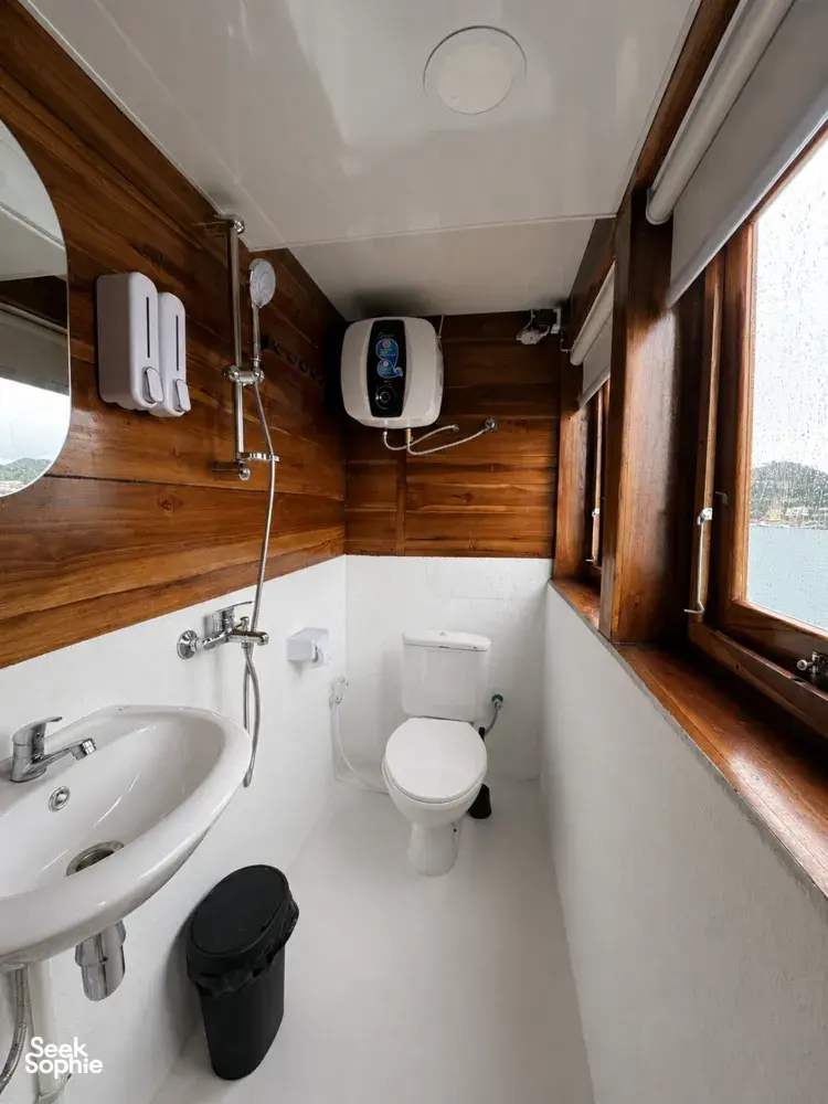 La Famille bathroom interior with a white toilet, wall-mounted sink and water heater, beside a window on a Komodo cruise Labuan Bajo