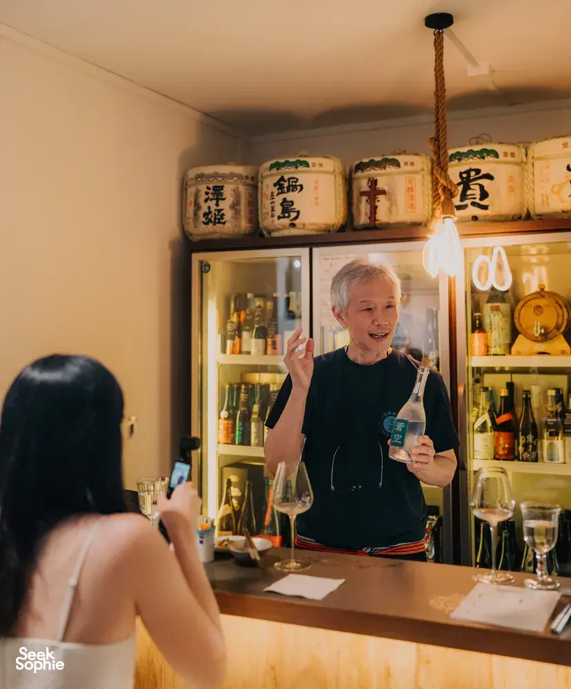 SakeMama pouring rare sake for guests during a Singapore tasting in a cosy shop, with glowing bottles and jars behind