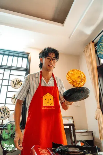 Host in red Jolie Cooking Class apron flips a round pancake while cooking at a home kitchen in Hoi An, Vietnam