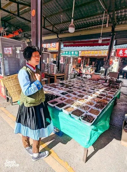 Fancy’s Yunnan cuisine host in Naxi dress browsing boxed herbs and mushrooms at a morning market stall in Lijiang