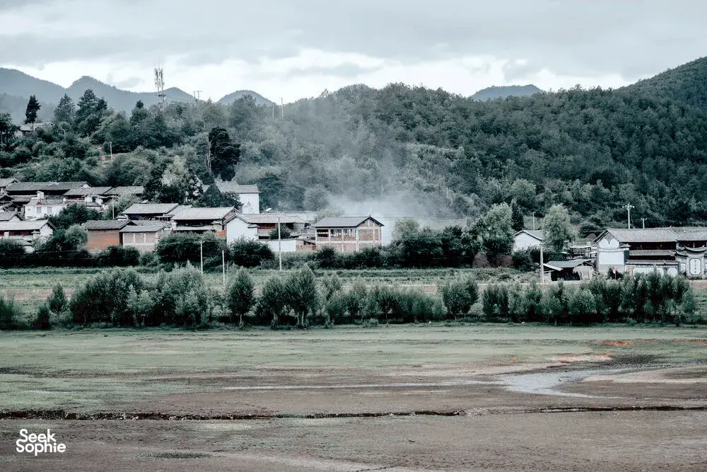 Pemandangan desa dari retret ramah lingkungan Linden Centre di Shaxi, Dali, dengan atap-atap halaman, hamparan ladang, dan pegunungan berkabut di balik pepohonan