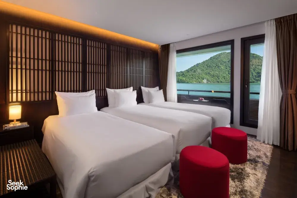 Oasis Bay Party Cruiser cabin with twin beds, red stools and curtains, with karst islands and bay water visible on the Halong Bay
