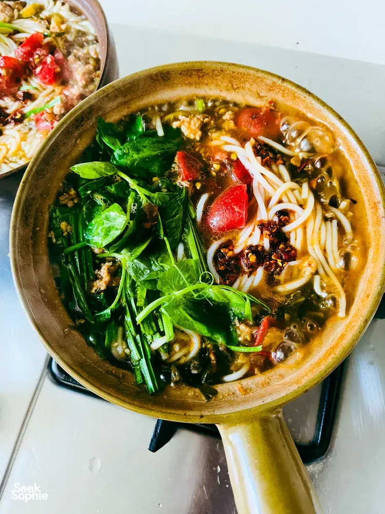 Bowl of steaming Yunnan-style noodle soup with greens, chilli and herbs on a table in Lijiang