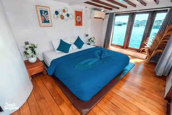 Phinisi cabin interior with a made-up double bed, blue bedding and sea-view windows for a relaxing La Famille Komodo cruise in Labuan Bajo