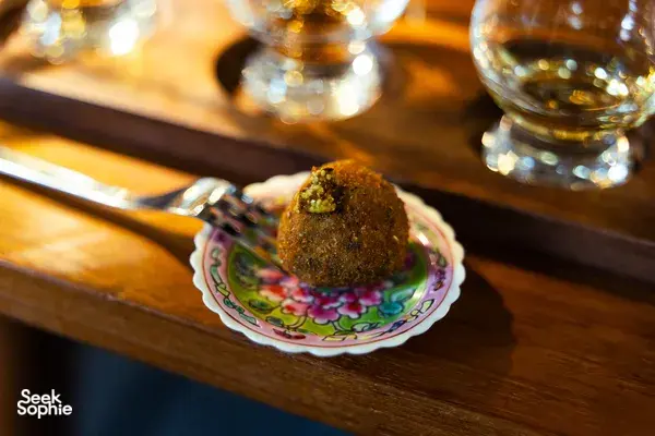 Decorative tasting plate with a small bite beside amber whisky glasses on a wooden bar, during a small-batch whisky tasting in Singapore
