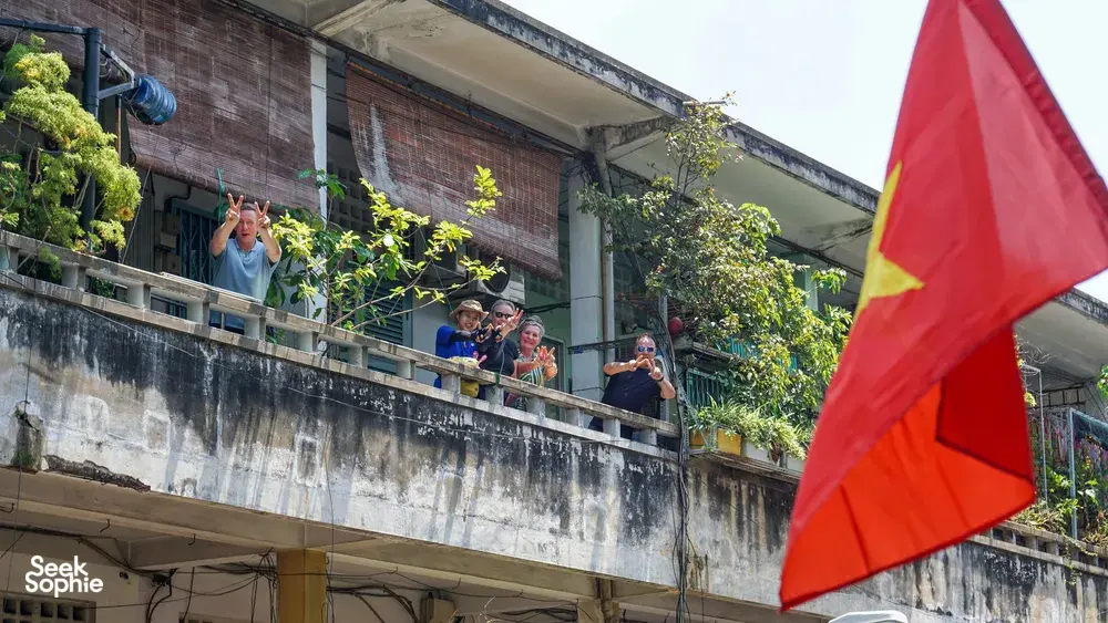 在胡志明市,一面越南国旗映满前景,一群旅行团游客在绿意环绕的高层阳台上微笑挥手