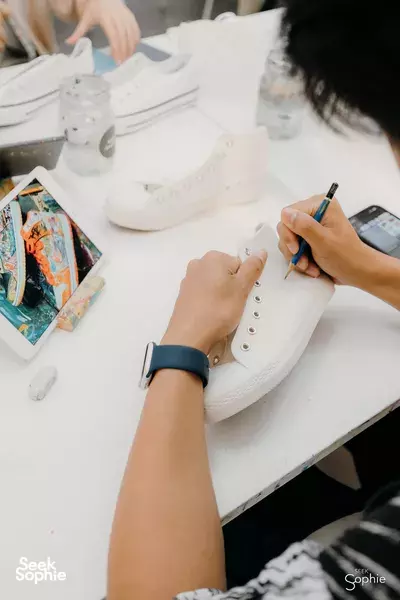 Hands brushing paint onto a white sneaker on a studio table, with paint bottles around, during Sneaker Art Jam workshops in Singapore