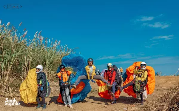 Group of paragliders in orange and yellow jackets and helmets posing beside a folded canopy on a hillside near Hanoi