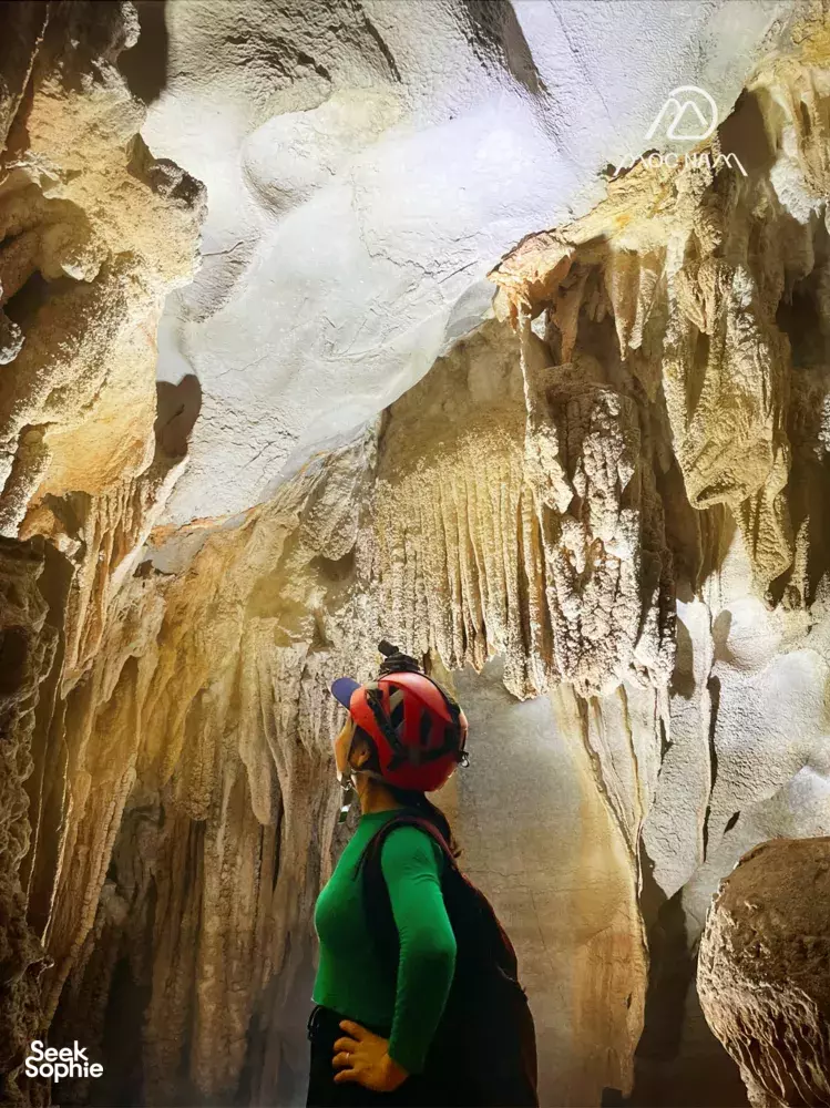 Caver in a green jacket and helmet looks up at illuminated stalactites inside a Phong Nha cave on a Moc Nam Adventure trek