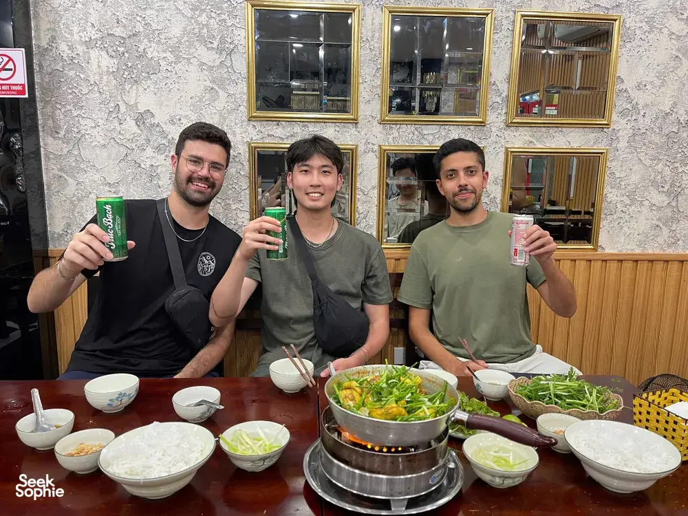 Travellers seated at a restaurant table with a shared hot pot and small bowls, enjoying a small-group Hanoi food adventure