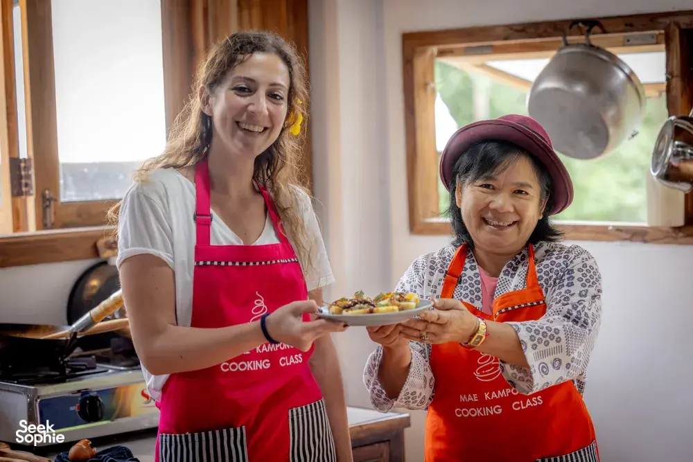 Two women in red aprons smiling while holding plated dishes at the end of the Mae Kampong forage-to-table cooking class in Chiang Mai
