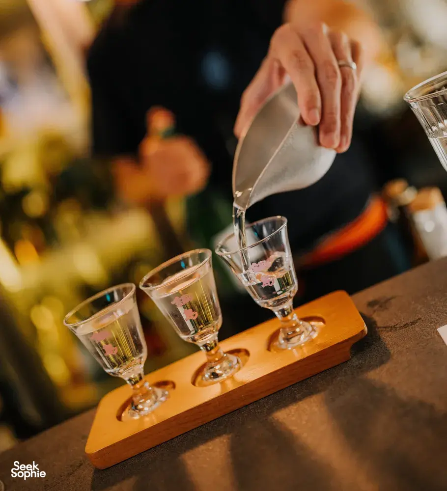 Pouring sake into small tasting glasses from a bottle at a rare Singapore session, with three glasses on a wooden tray