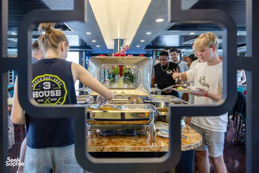 Travellers gather at a buffet counter with hot dishes on a Halong Bay party cruise, chatting in a bright dining room