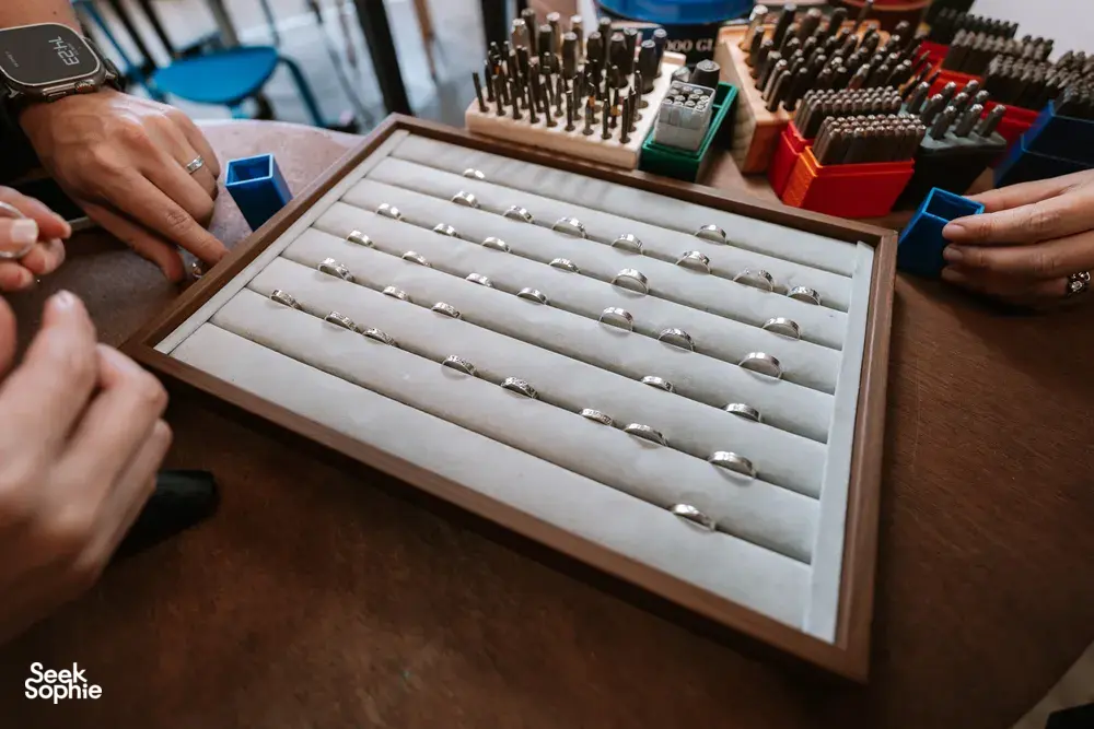 Open wooden display tray filled with silver ring bands as hands choose pieces at Fat Anvil Studios in Singapore couple ring workshop