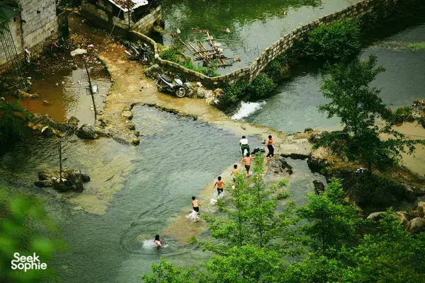 People wading in a shallow clear river in Cao Bang, with motorbikes parked along stone embankments and a relaxing crowd