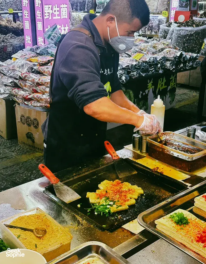 Local cook at a Lijiang street-food stall spreading savoury batter on a sizzling griddle with chopped greens and sauce