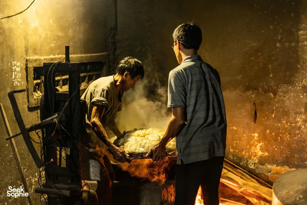 Blacksmiths working by a blazing forge in Hoi An, one man holding hot metal while another turns tools near the fire