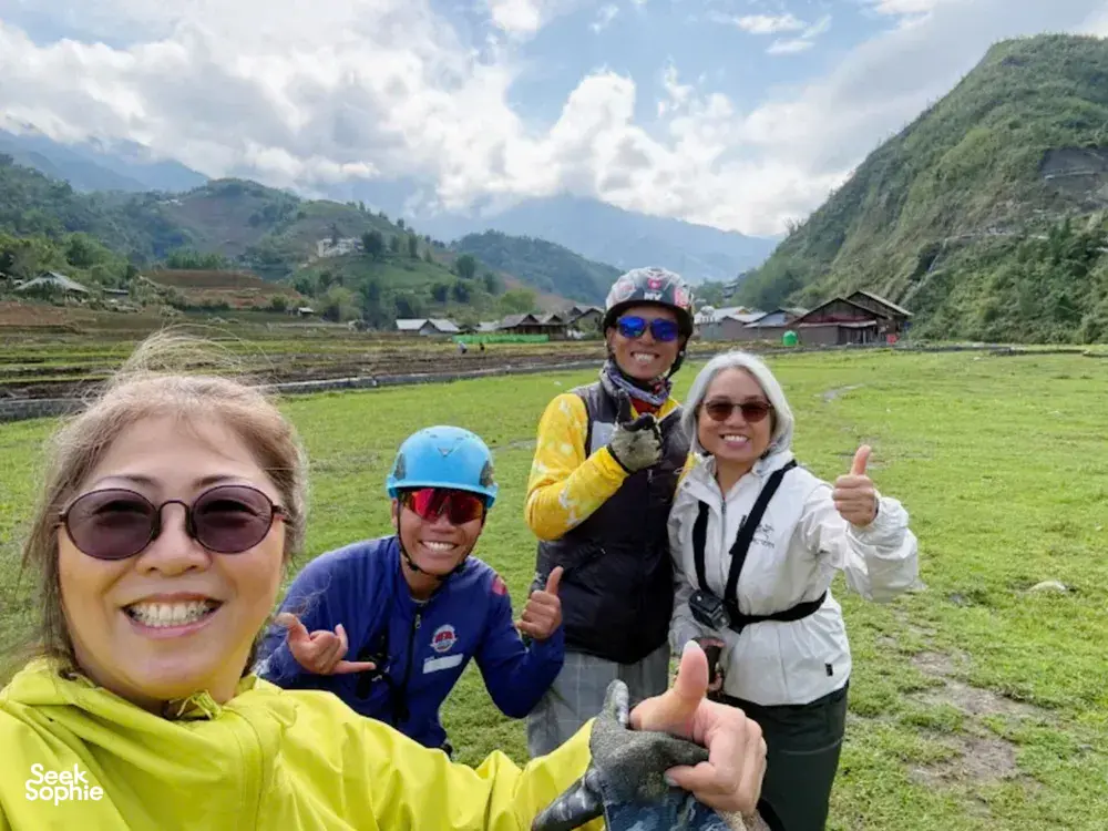 Group of paragliding passengers in helmets smiling and giving thumbs up on a grassy launch field in the hills, Sapa